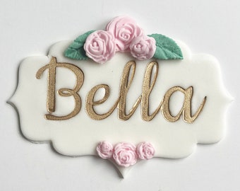 May include: White cake topper with the name "Bella" written in gold script. The topper is decorated with pink and green sugar flowers.