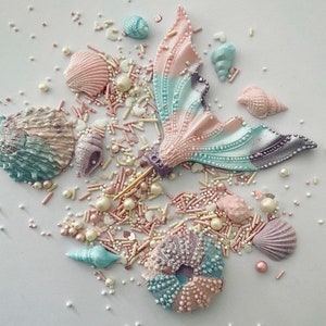 May include: A pastel blue and pink mermaid tail cake topper with a gold stick, surrounded by pastel blue, pink, and white sprinkles and pearl-like decorations. The sprinkles are in the shape of seashells, stars, and hearts.