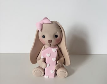 May include: A beige bunny figurine with a pink bow and a pink number one. The number one is decorated with white hearts. The bunny has black eyes and is sitting down.