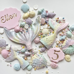 May include: A set of mermaid tail and seashell cake toppers with sprinkles and a pink cake topper with the name "Ellen" and the number "5" in gold lettering.