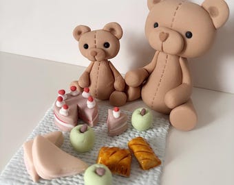 Op de afbeelding: Twee bruine teddybeerbeeldjes zitten in de buurt van een miniatuur picknickscène. Een taart met rode bessen, stukjes taart, groene appels en gebak liggen op een witte, getextureerde mat. De beren hebben gestikte details.