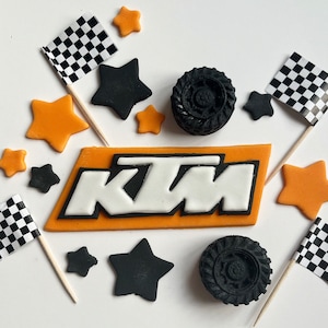 May include: Orange and white edible cake topper with the letters "KTM" in white with black outlines. The topper is surrounded by black and white chequered flags, orange stars, and black tyre-shaped decorations.