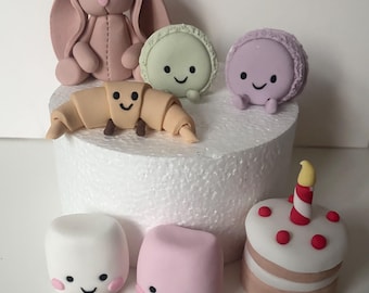 Op de afbeelding: Een verzameling kleifiguurtjes met een oudroze konijn, een lichtgroene macaron, een lavendelkleurige macaron, een beige croissant, een witte marshmallow, een roze marshmallow en een klein taartje met een rood-witte kaars, allemaal met gezichtjes.