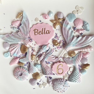 May include: A collection of edible mermaid-themed cake decorations including four iridescent pastel mermaid tails, various seashells, starfish, and two pink scalloped plaques. One plaque features the name Bella and the other the number 6.