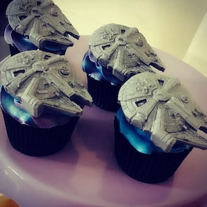 May include: Four cupcakes decorated with grey fondant Millennium Falcon spaceship toppers. The cupcakes have black liners and blue and purple frosting.