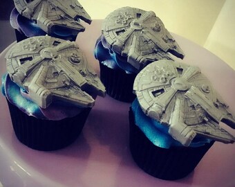 May include: Four cupcakes decorated with gray fondant Millennium Falcon spaceship toppers. The cupcakes have black liners and blue and purple frosting.