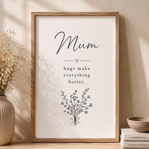 Op de afbeelding: Een ingelijste verticale wandprint met het woord Mum in een scriptlettertype, gevolgd door een klein hartpictogram en de tekst hugs make everything better. Onder de tekst staat een zwarte lijntekening van een boeket wilde bloemen op wit papier.