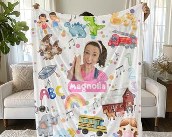 May include: A white fleece blanket with a colorful design featuring a school bus, a rainbow, a farm scene, a girl with a bubble, a dinosaur, a boat, bunnies, an elephant, musical notes, letters, and numbers. The blanket also has a pink rectangle with the name "Magnolia" written in white.