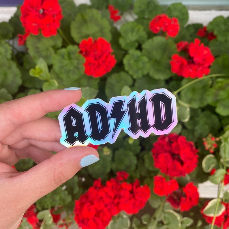 Pu&ograve; includere: Un adesivo olografico con la parola "ADHD" in lettere maiuscole nere con un fulmine al centro. L'adesivo &egrave; su una mano su uno sfondo di fiori rossi.