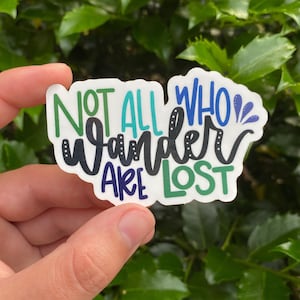 Può includere: Un adesivo bianco con il testo "Not all who wander are lost" in lettere nere, verdi, blu e turchese. L'adesivo ha una linea punteggiata attorno al testo.