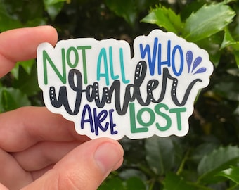 Peut inclure: Un autocollant blanc avec le texte "Not all who wander are lost" en lettres noires, vertes, bleues et turquoise. L'autocollant a une ligne pointillée autour du texte.