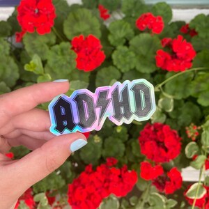 Pu&ograve; includere: Un adesivo olografico con la parola "ADHD" in lettere nere con un fulmine al centro. L'adesivo &egrave; su una mano su uno sfondo di fiori rossi.