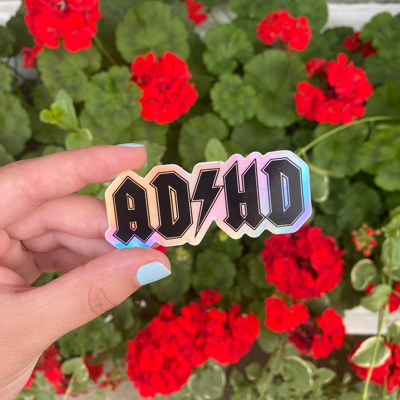 Pu&ograve; includere: Un adesivo olografico con il testo "ADHD" in lettere maiuscole nere con un fulmine tra la "D" e la "H".