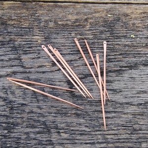 May include: A set of ten copper needles, scattered on a wooden surface. The needles are long and thin, with a sharp point at one end and a small eye at the other.