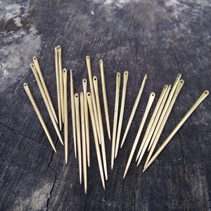 May include: A collection of 20 gold-colored sewing needles arranged in a fan shape on a dark wood surface.
