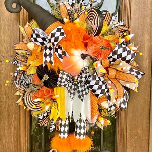 May include: A Halloween-themed wreath with a witch design. It features a black witch hat, orange and black checkered bows, and decorative elements like pumpkins and candy corn. The word "Alluring" is displayed at the bottom.