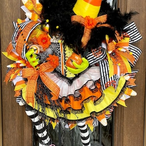 May include: A Halloween-themed wreath with a candy corn hat, black feathers, and a witch's dress design. The wreath features orange, yellow, and black ribbons, a green hand holding a pumpkin, and striped legs. The text "Alluring Wreath Designs" is at the bottom.