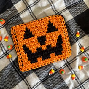May include: An orange and black crocheted coaster in the shape of a jack-o'-lantern face. The coaster is square with a black border and is surrounded by Halloween candy corn and pumpkin candies on a black and white plaid background.