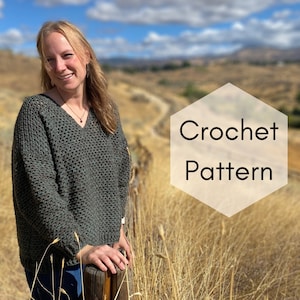 May include: A woman wearing a dark green crochet sweater with a V-neck. The image includes the text "Crochet Pattern" in a white hexagon shape. The background features a field of dry grass and a blue sky.