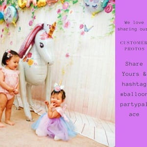 May include: Two young children pose with a large unicorn balloon. One child wears a pink outfit, the other a blue and purple dress with a unicorn horn headband. The background features floral decorations and text that reads "We love sharing our customer photos."