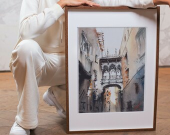 May include: A framed watercolor painting of a European street scene. The artwork features a bridge and buildings in shades of brown, gray, and blue. The frame is a dark wood with a white mat.
