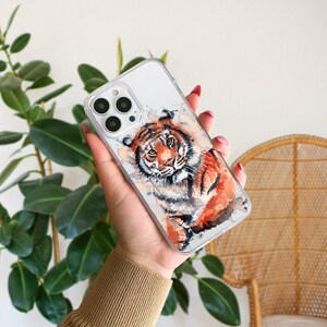 May include: A clear phone case with a watercolor illustration of a tiger. The tiger is orange and black with white stripes. The case is designed for an iPhone.