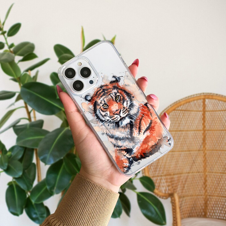May include: A clear phone case with a watercolor illustration of a tiger. The tiger is orange and black with white stripes. The case is designed for an iPhone.