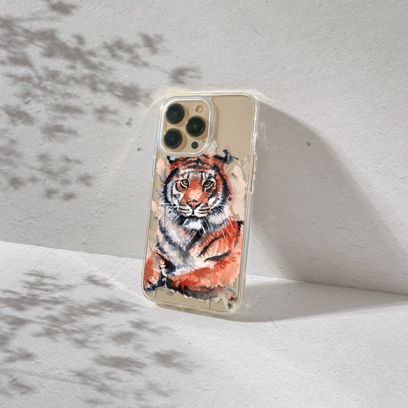 May include: A clear phone case with a tiger illustration. The tiger is orange, black, and white with a detailed watercolor style. The phone case has a gold trim around the camera lens.