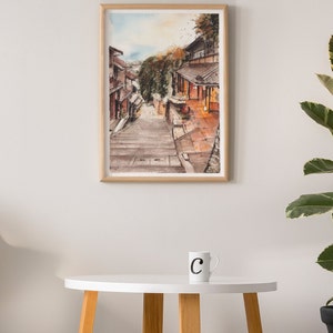 May include: A watercolor painting of a narrow street in a Japanese village. The street is lined with traditional wooden buildings and leads up a set of stone steps. The painting is framed in a light wood frame.