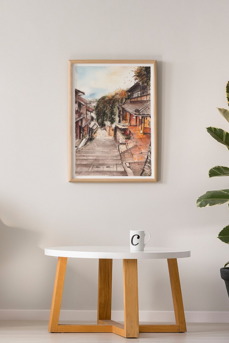 May include: A watercolor painting of a narrow street in a Japanese village. The street is lined with traditional wooden buildings and leads up a set of stone steps. The painting is framed in a light wood frame.