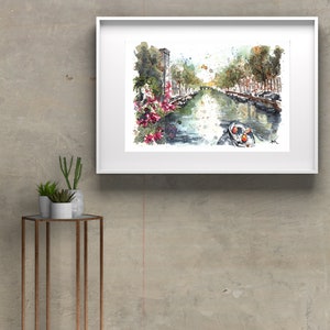 May include: Watercolor painting of a canal in a city with buildings on either side. The canal is lined with trees and there is a small boat in the water. The painting is framed in a white frame.