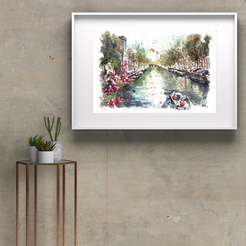 May include: Watercolor painting of a canal in a city with buildings on either side. The canal is lined with trees and there is a small boat in the water. The painting is framed in a white frame.