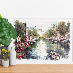 May include: Watercolor painting of a canal in a city with buildings on either side. The canal is filled with water and there are boats in the water. There are trees on either side of the canal and flowers in the foreground. The painting is framed in a white frame.