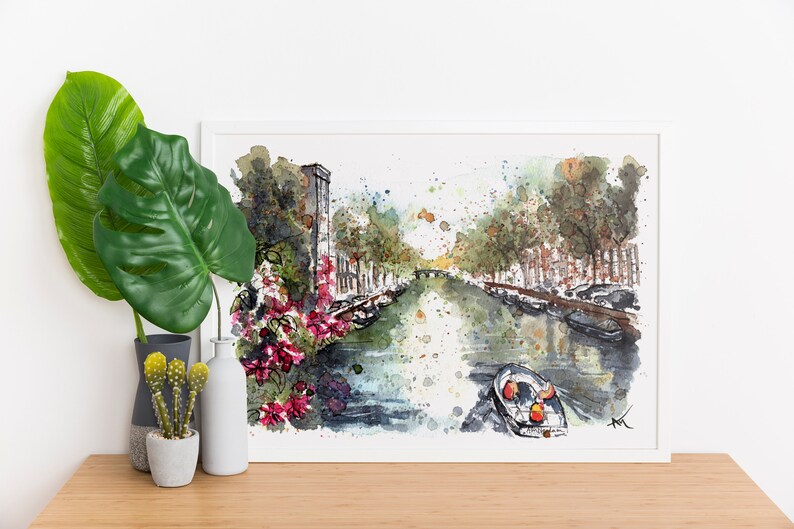 May include: Watercolor painting of a canal in a city with buildings on either side. The canal is filled with water and there are boats in the water. There are trees on either side of the canal and flowers in the foreground. The painting is framed in a white frame.