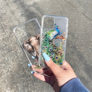 May include: Two clear phone cases with different designs. One case features a peacock with blue, green, and gold accents. The other case features an elephant with brown and gray accents.