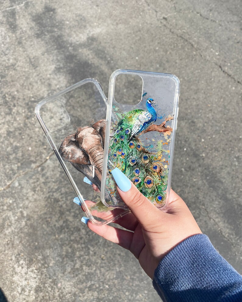 May include: Two clear phone cases with different designs. One case features a peacock with blue, green, and gold accents. The other case features an elephant with brown and gray accents.
