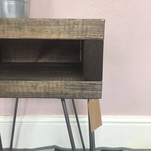 May include: A small, rustic wooden side table with a dark stained finish and a lower shelf. The table has metal hairpin legs and a small tag attached. The wood has visible grain and a distressed appearance.