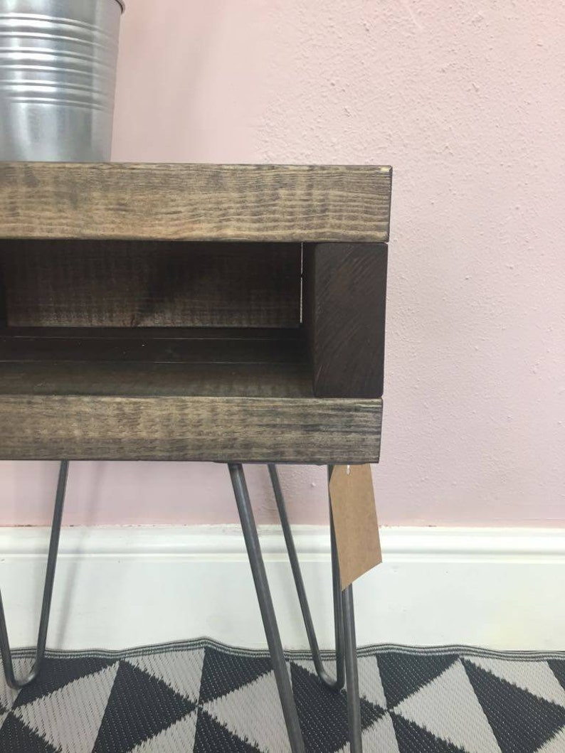 May include: A small, rustic wooden side table with a dark stained finish and a lower shelf. The table has metal hairpin legs and a small tag attached. The wood has visible grain and a distressed appearance.