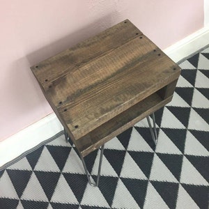 May include: A small, rustic wooden side table with a dark brown finish. The table has a lower shelf and is supported by four silver hairpin legs. The table is placed on a black and white geometric patterned rug.