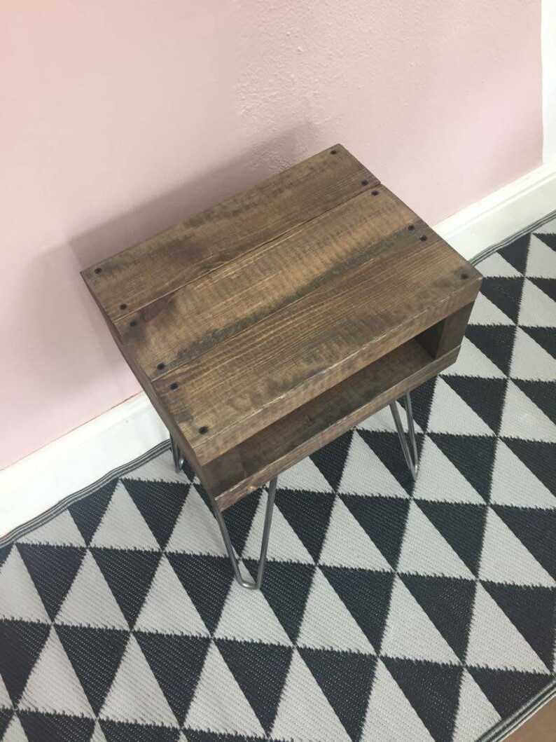 May include: A small, rustic wooden side table with a dark brown finish. The table has a lower shelf and is supported by four silver hairpin legs. The table is placed on a black and white geometric patterned rug.