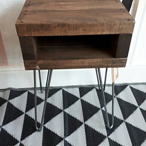 May include: A small, rustic wooden side table with a dark brown finish. The table features a rectangular top with a lower shelf and is supported by four metal hairpin legs. The table measures approximately 20 inches in height.
