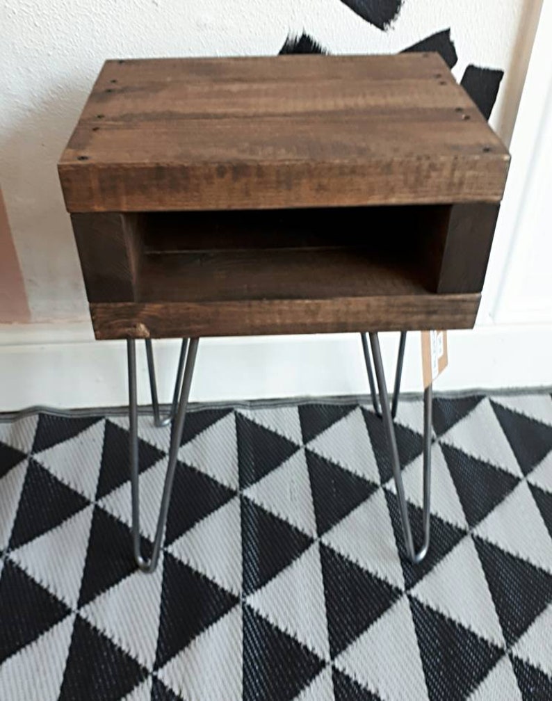 May include: A small, rustic wooden side table with a dark brown finish. The table features a rectangular top with a lower shelf and is supported by four metal hairpin legs. The table measures approximately 20 inches in height.