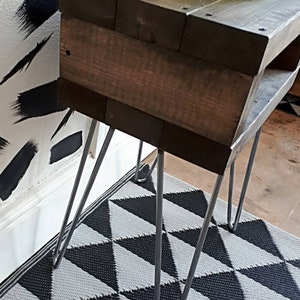 May include: A small, rustic wooden side table with dark brown stain. The table has a rectangular top and a lower shelf, supported by silver hairpin legs. A decorative vase sits on top. The table is placed on a black and white geometric patterned rug.