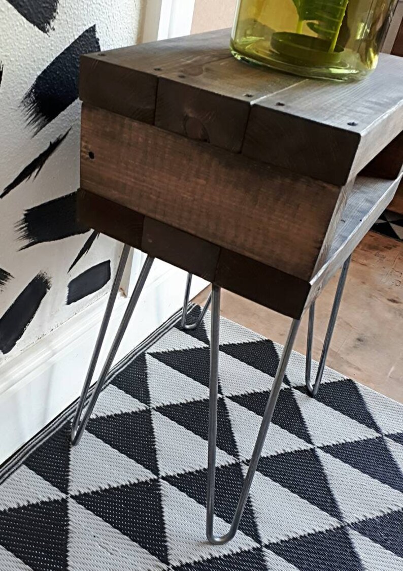 May include: A small, rustic wooden side table with dark brown stain. The table has a rectangular top and a lower shelf, supported by silver hairpin legs. A decorative vase sits on top. The table is placed on a black and white geometric patterned rug.