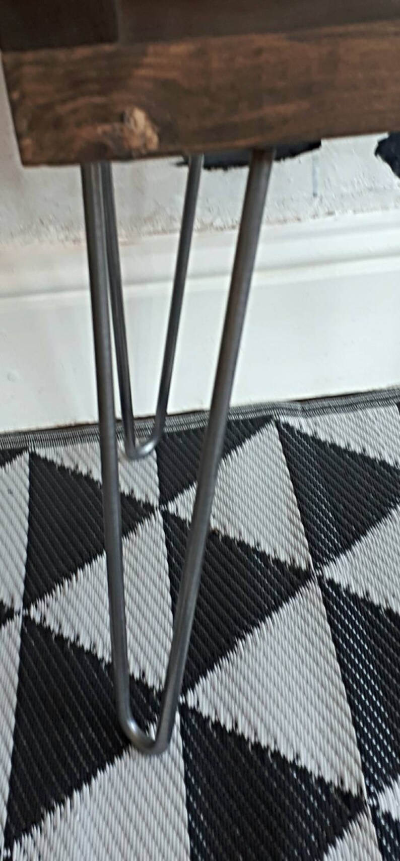 May include: Close-up of a wooden table with metal hairpin legs. The tabletop is a dark brown color, and the legs are silver. The table is set on a black and white geometric patterned rug.