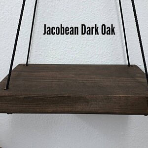 May include: A dark brown, rectangular wooden shelf suspended by black rope. The shelf is stained with a dark oak finish and has rounded edges. The text "Jacobean Dark Oak" is displayed above the shelf.