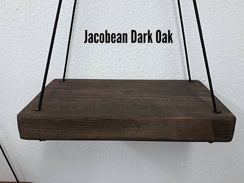 May include: A dark brown, rectangular wooden shelf suspended by black rope. The shelf is stained with a dark oak finish and has rounded edges. The text "Jacobean Dark Oak" is displayed above the shelf.