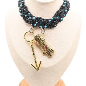 May include: A black braided cord choker necklace featuring small embedded turquoise beads. Two gold-toned metal charms hang from the center: a detailed gauntlet with a red stone and a long, slender arrow-shaped pendant inscribed with the text "Sania Corona".
