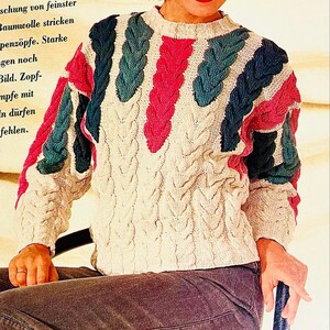 Könnte beinhalten: Ein weißer Strickpullover mit einem Zopfmuster. Der Pullover hat rosa und blaugrüne geflochtene Streifen über der Brust und den Schultern. Der Text "für Wandervögel: Aus schung von feinster Baumwolle stricken penzöpfe. Starke gen noch Bild. Zopf-mpfe mit In dürfen fehlen." ist auf der linken Seite des Bildes sichtbar.
