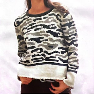 Könnte beinhalten: Ein langärmliger Pullover mit einem Camouflage-Muster in Grau-, Schwarz- und Weißtönen. Der Pullover hat einen Rundhalsausschnitt und einen weißen Saum und Bündchen. Das Design ist ein sich wiederholendes geometrisches Muster.